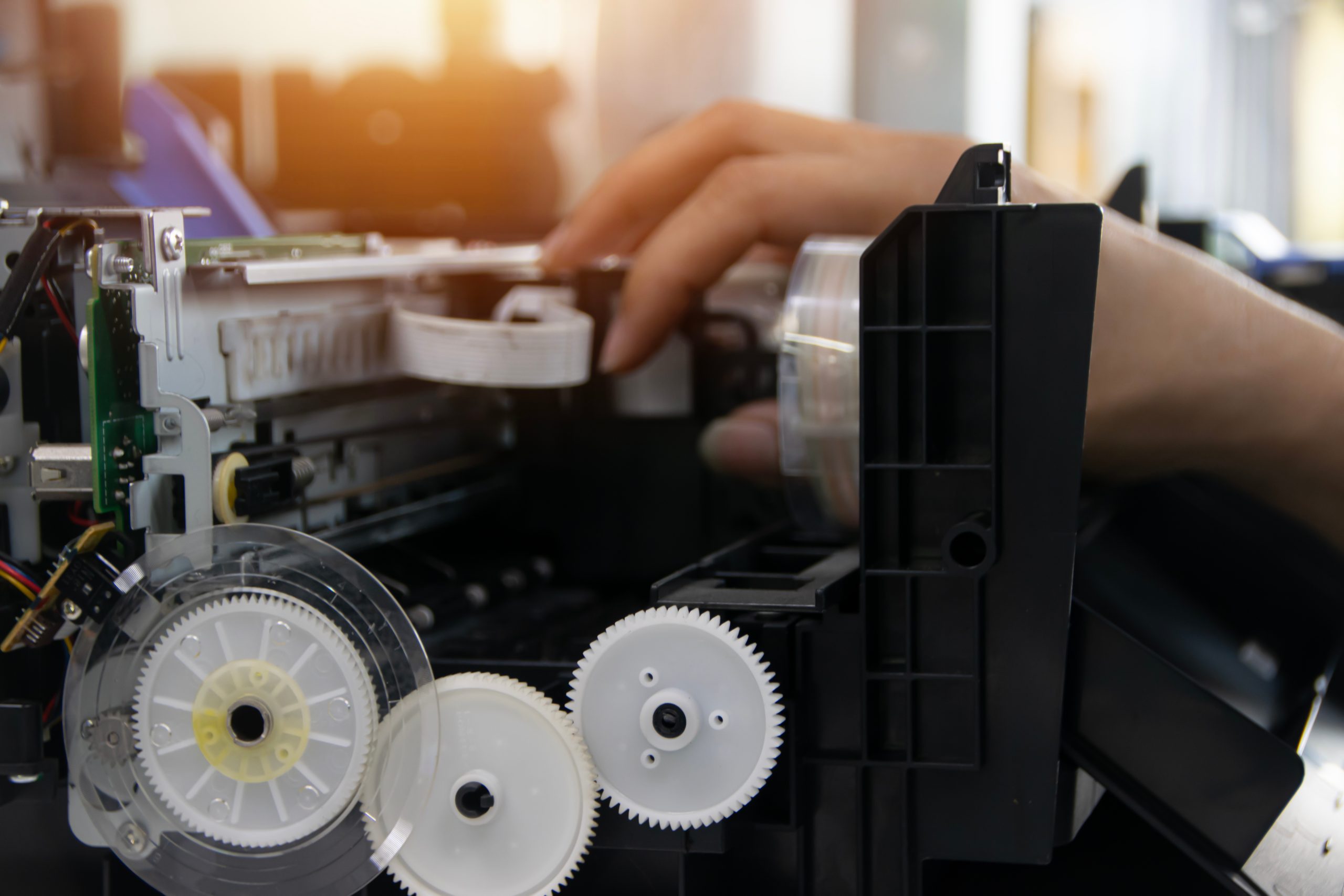 Technician's hands servicing a printer's internal components and replacement parts during repair work.